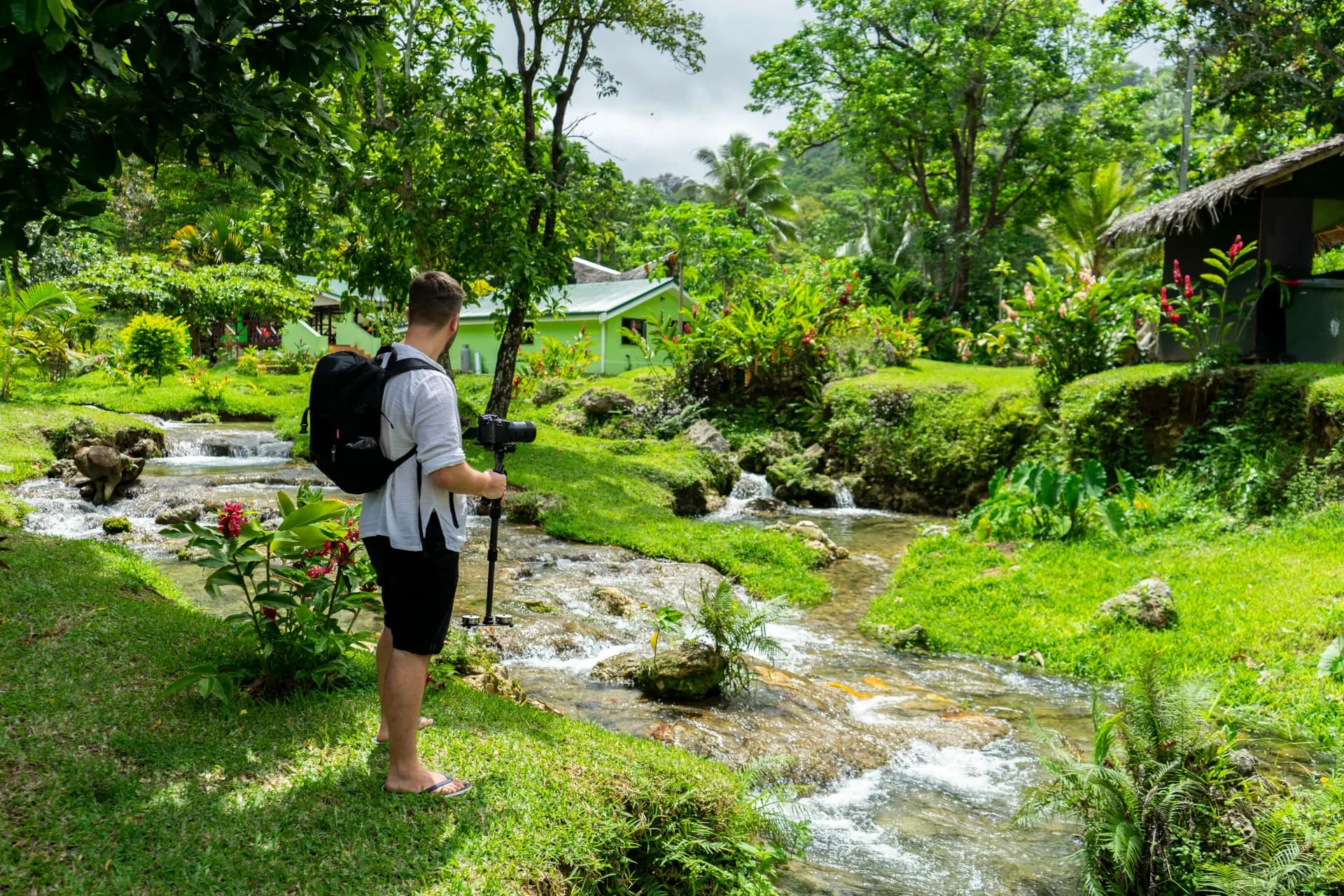 man in grey shirt and black shorts standing by the river bank near house during daytime