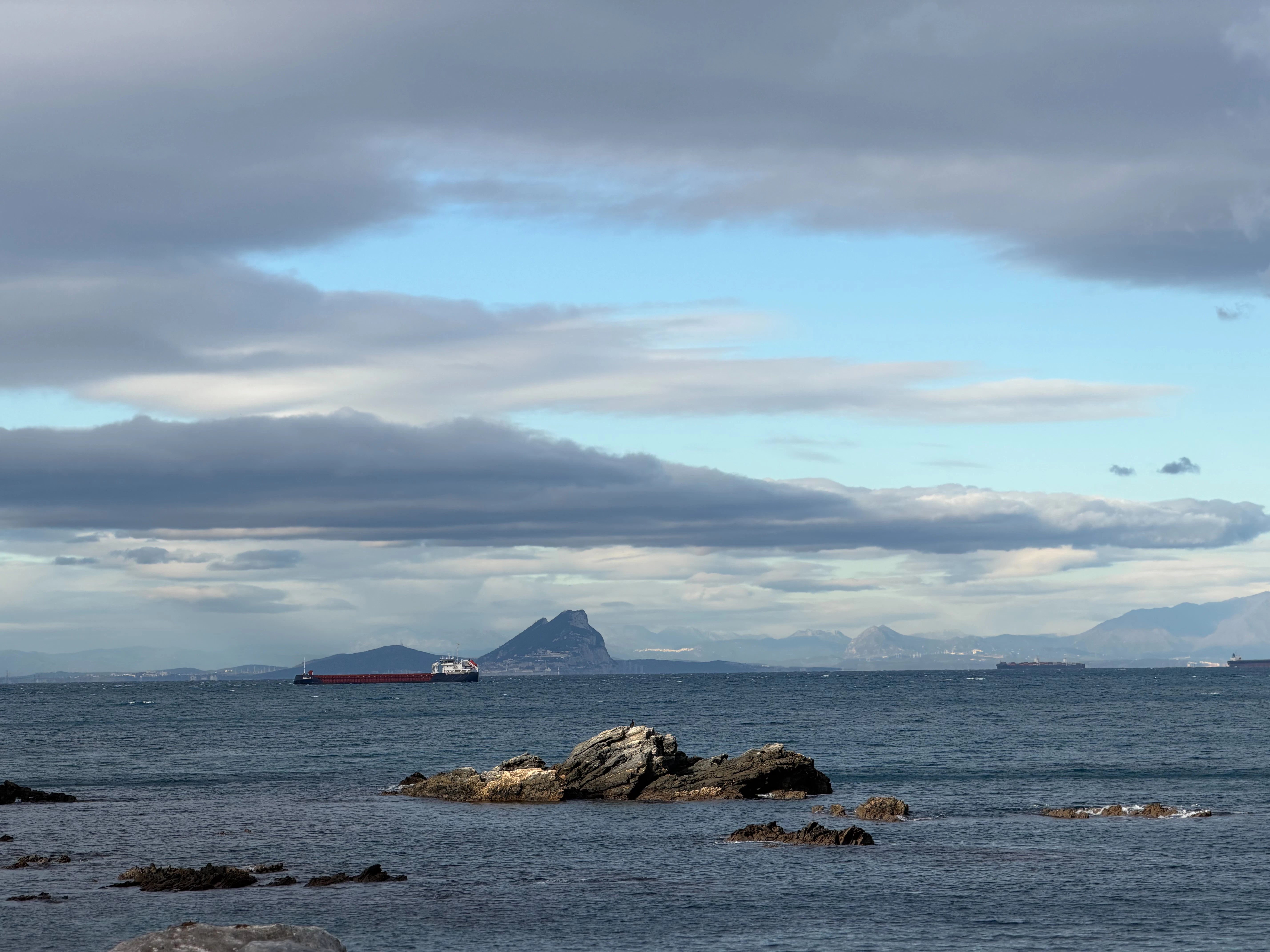 A cargo ship sails past a rocky outcrop at sea.