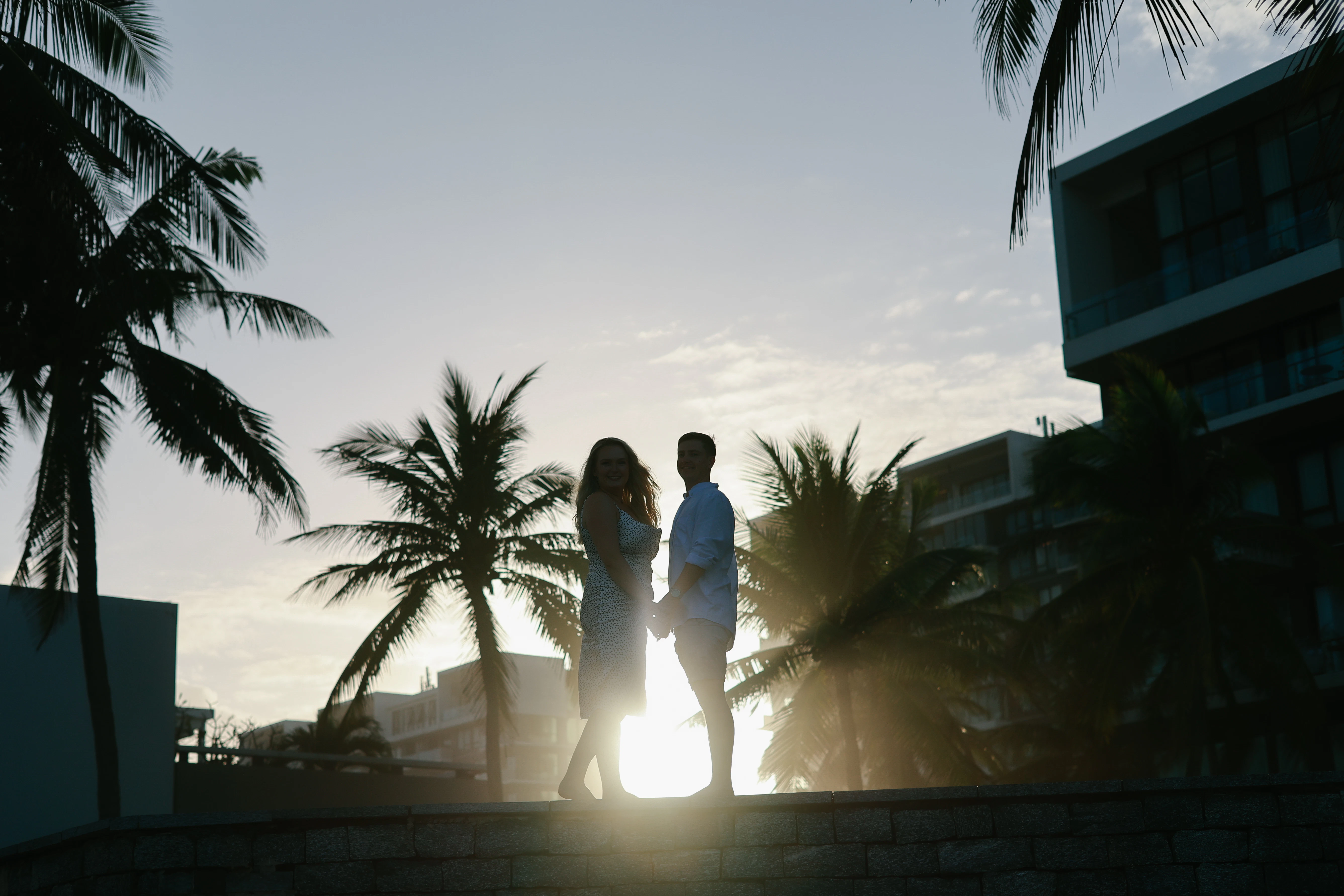 Couple holding hands against a sunset with palm trees.