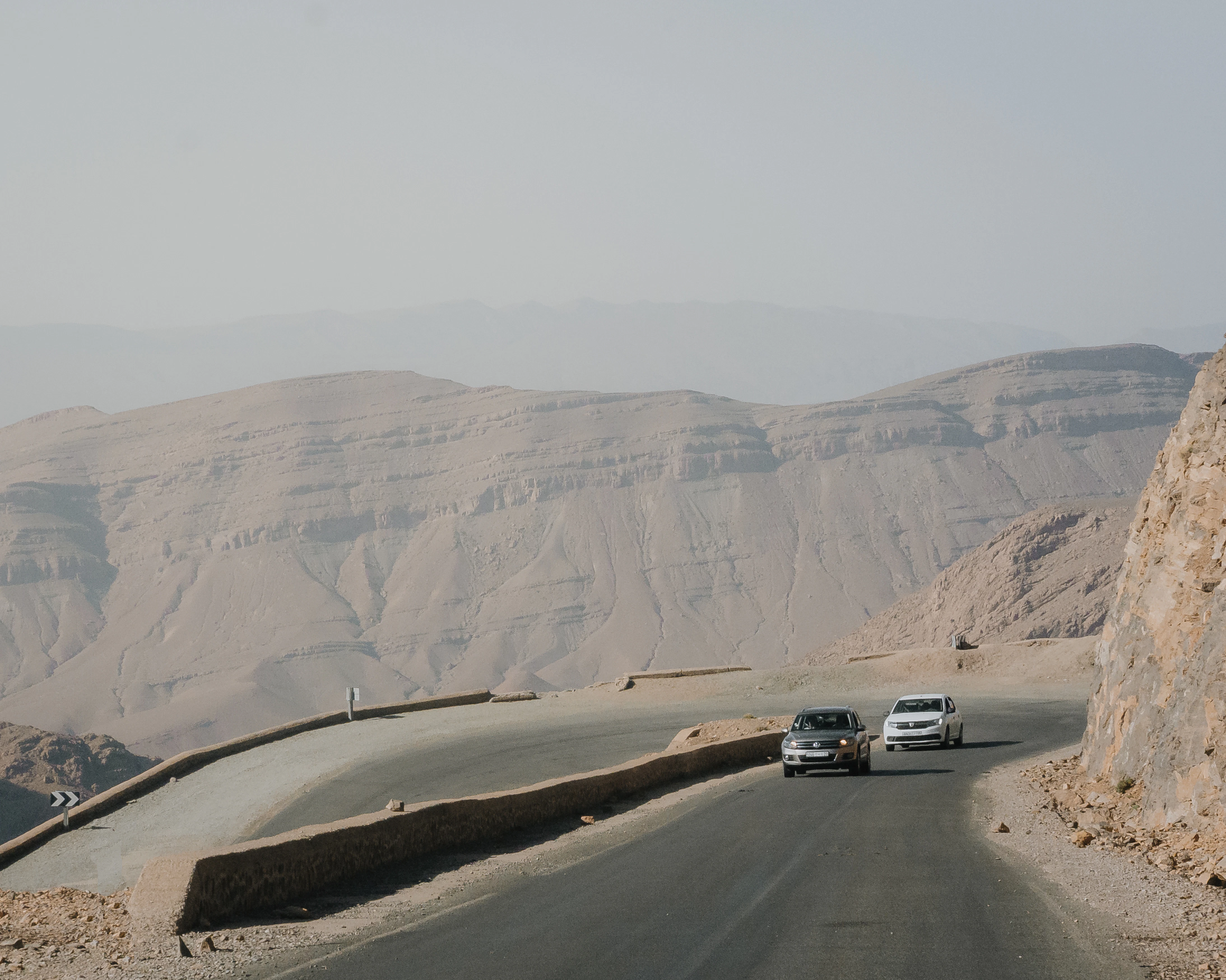 Two cars drive on a winding road through desert mountains.